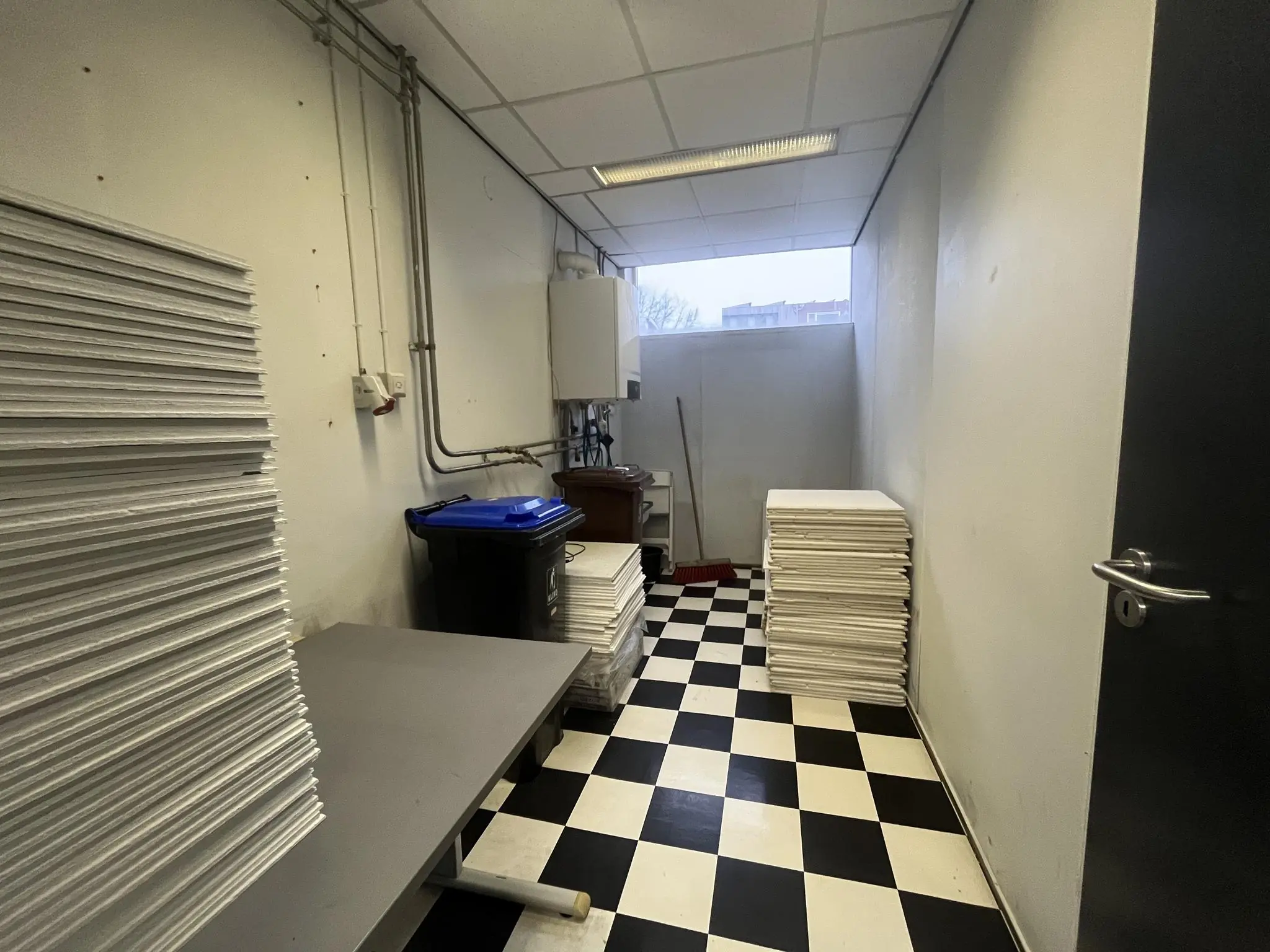 Opslagruimte met zwart-wit geblokte vloer, grote stapels witte panelen en schoonmaakmaterialen aan de Burgemeester Smitsstraat.